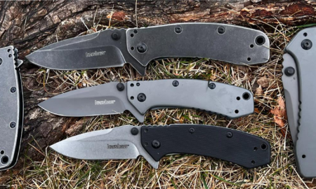 Three Kershaw folding pocket knives displayed outdoors on grass near a log.