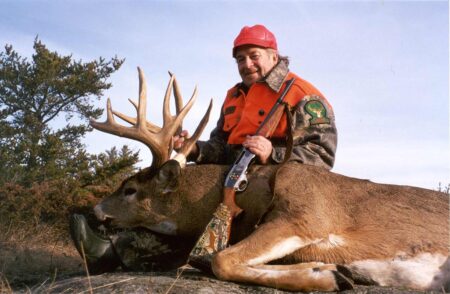 6 Best Deer Hunting Rifles for The Big Woods Hunter in blaze orange poses with large whitetail buck and rifle outdoors.