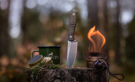 Fixed blade camping knife balanced on log stump beside open flame and mug.