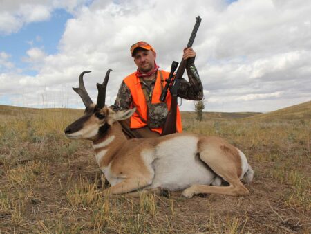 Hunter in orange vest poses with harvested pronghorn antelope and rifle outdoors.