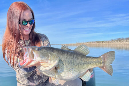 Woman angler proudly holding a large trophy largemouth bass on a lake.