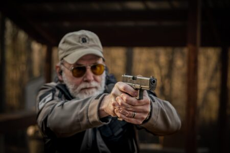 Older man with white beard aiming pistol at outdoor shooting range
