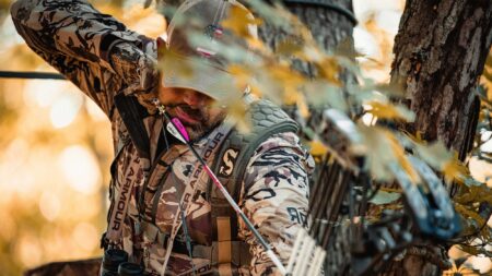 Hunter in camouflage Under Armour gear holding bow in autumn forest tree stand