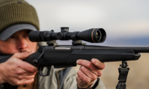 Person aiming bolt-action rifle with scope mounted on bipod outdoors