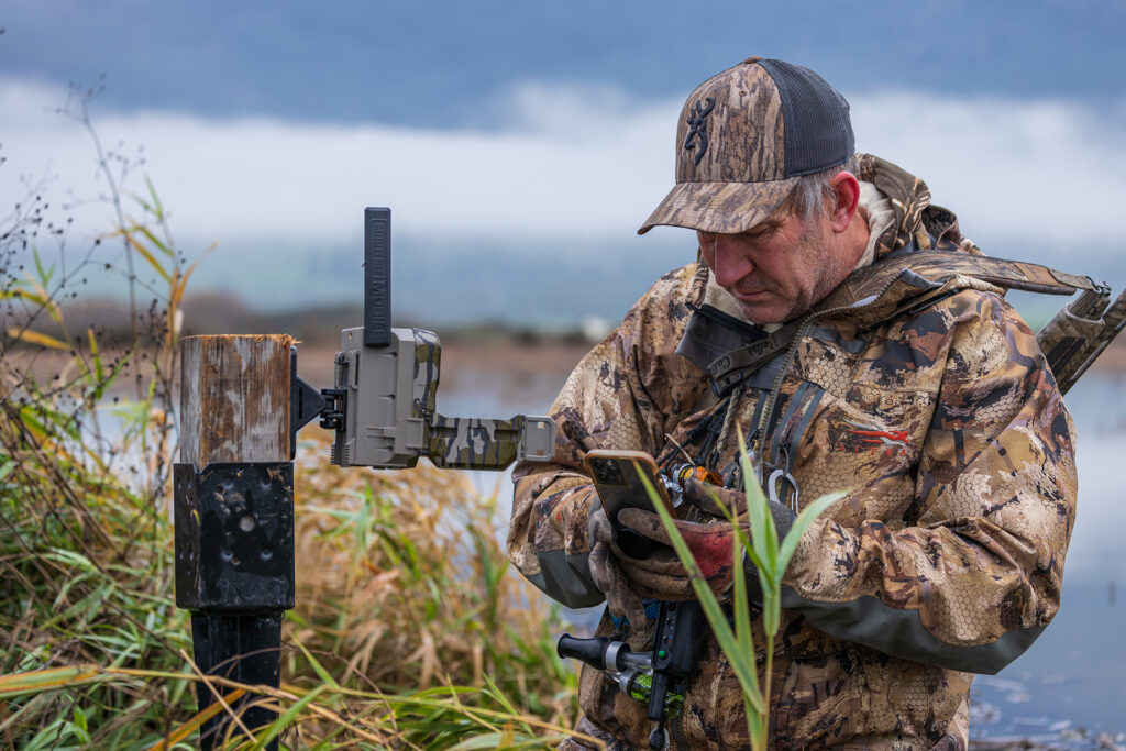 Hunter in camouflage gear checking trail camera near wetland with tablet device.