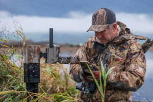 Hunter in camouflage gear checking trail camera near wetland with tablet device.