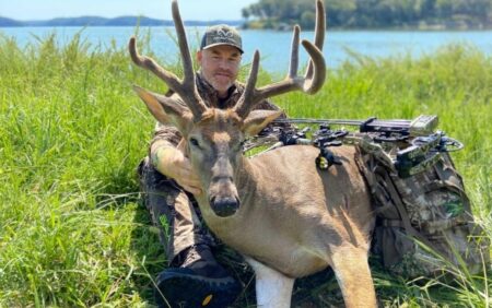 Hunter in camo poses with harvested whitetail buck and compound bow outdoors.