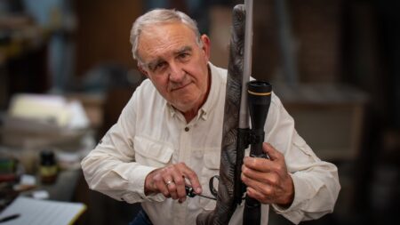 Elderly man holding a scoped hunting rifle in an indoor workshop setting.