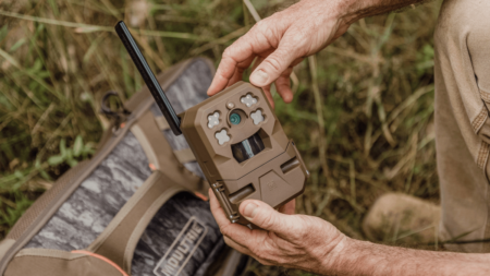 Person holding a wireless trail camera outdoors near hunting gear in grass