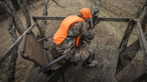 Hunter in blaze orange vest aiming rifle from elevated wooden deer stand