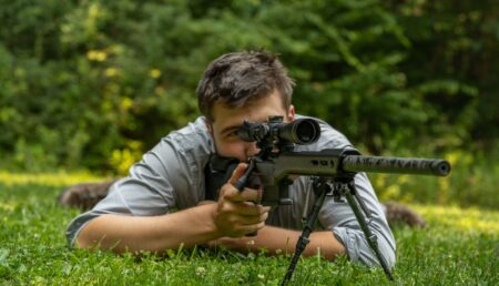 Young man lying in grass aiming precision rifle with scope and bipod