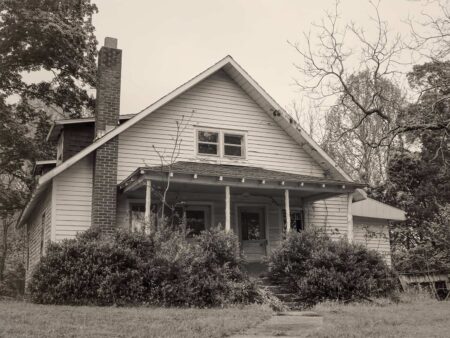 Giving Up the Ghosts Abandoned white craftsman bungalow with brick chimney surrounded by overgrown shrubs and trees