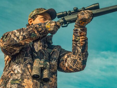 Hunter in camouflage aiming scoped rifle with Nikon binoculars hanging from neck