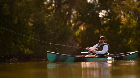 Man fly fishing from green canoe on calm river surrounded by trees