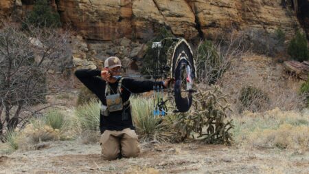 Archer kneeling in desert canyon aiming compound bow among cacti and red rocks