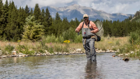 Man fly fishing in mountain stream wearing waders and holding fishing net.