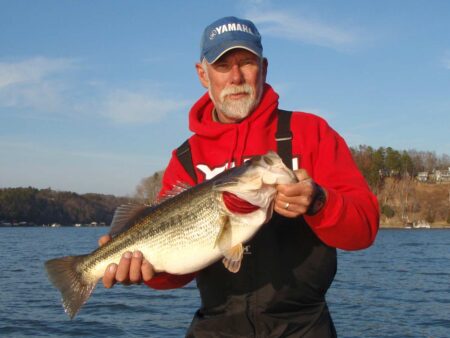 Man in red Yamaha hoodie proudly holds large largemouth bass on lake.