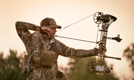 Hunter in camouflage aiming compound bow at sunset during archery hunting season
