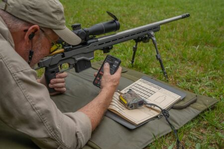 Shooter in prone position using smartphone ballistics app beside scoped rifle on bipod