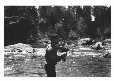 Man fishing with rod in rocky river surrounded by trees, black and white photo