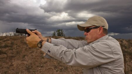 How to Hold a Pistol—the Right Way Man in cap and sunglasses aiming pistol outdoors under dramatic storm clouds.