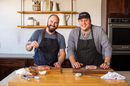 Behind the Brand: An Interview with the Founders of Porter Road Two smiling chefs preparing raw steaks on wooden cutting boards in a kitchen.