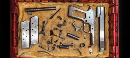 Disassembled 1911 pistol parts laid out in a red plastic crate on burlap