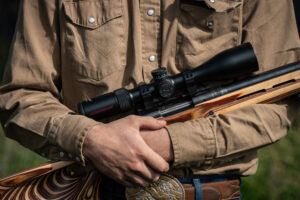 Person holding bolt-action rifle with scope and laminate wood stock outdoors.