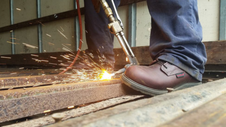Worker using welding torch on metal surface with sparks flying near safety boots.