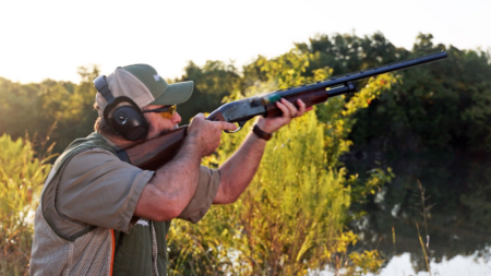 Man wearing ear protection aims shotgun outdoors near water and trees.