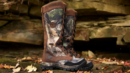 Tall camouflage hunting boots with brown leather and lug soles on rocky ground