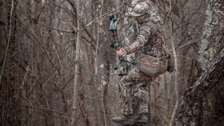 Saddle Hunting 101: A Beginner’s Guide Camouflaged bowhunter standing in tree stand holding compound bow in winter forest.