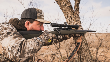 Hunter in camouflage aiming bolt-action rifle with scope outdoors near tree
