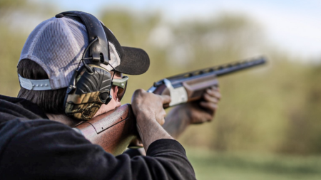 Person wearing ear protection aiming shotgun outdoors during clay pigeon shooting sport