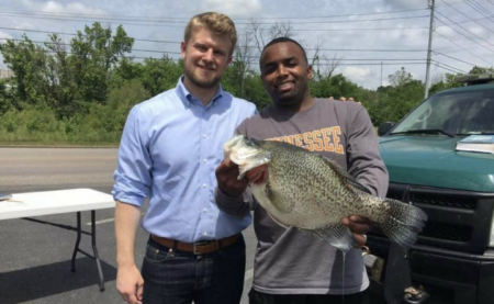 Two men smiling outdoors holding a large crappie fish near a parking lot.