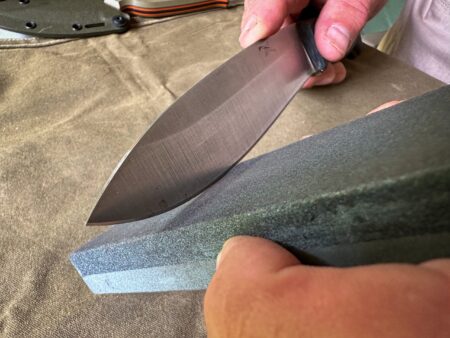Person sharpening a chef's knife on a whetstone sharpening block outdoors.