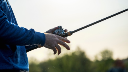 Person holding fishing rod with baitcasting reel outdoors at dusk near water