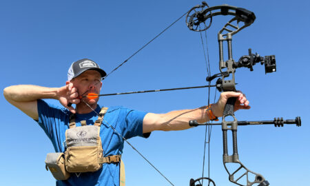 Man aiming compound bow with orange arrow against clear blue sky outdoors
