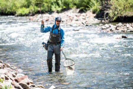 Fly fisherman wading in rocky mountain river holding net and fishing rod