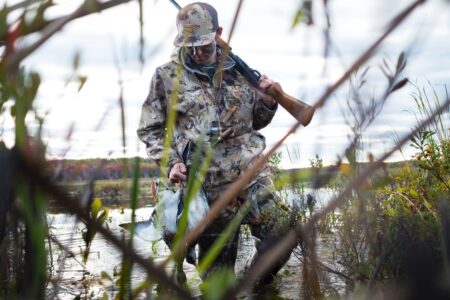The Best Duck Hunting Jackets of 2024, Tested and Reviewed Hunter in camouflage carrying shotgun and harvested duck through wetland marsh.