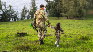 The Best Father’s Day Hunting Gifts for Dad Adult hunter in camouflage holds child's hand while carrying compound bow outdoors.