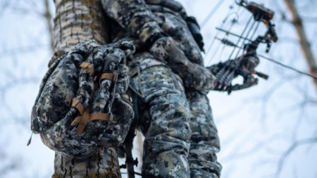 Camouflaged bowhunter climbing snow-covered tree with compound bow in winter forest