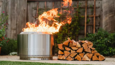 Best Solo Stove Deals of 2024 Solo Stove stainless steel fire pit burning with flames beside stacked firewood outdoors