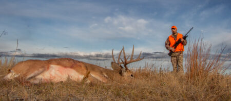 Hunter in orange vest stands beside harvested whitetail buck in open field.