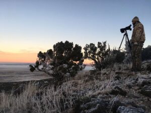 Hunter in camouflage using spotting scope on tripod at dawn in desert landscape.
