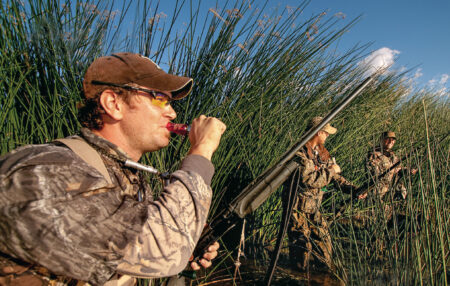 Three hunters in camouflage use duck calls and shotguns in marsh reeds.