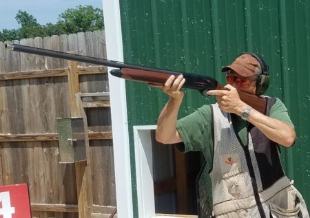 Man in shooting vest aims shotgun at clay pigeon shooting range outdoors.