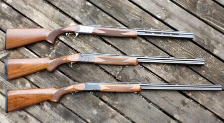 Three wooden stock shotguns with blue steel barrels displayed on weathered deck boards.