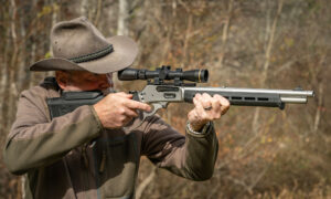 Man in cowboy hat aiming scoped lever-action rifle outdoors in wooded setting
