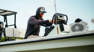 Man with beard steering a fishing boat wearing sunglasses and hoodie.